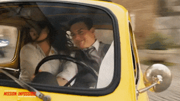 Movie gif. Tom Cruise as Ethan Hunt in "Mission Impossible: Dead Reckoning," drives a yellow Fiat 500 through a European city, screeching around corners. Haley Atwell as Grace sits in the passenger seat, whirling her head around to look behind them with an alert expression. 