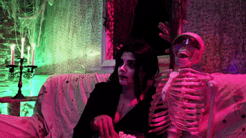 Video gif. Woman wearing a vampire costume eats popcorn on a couch next to a skeleton wearing sunglasses as a creepy monster emerges from the window behind them and waves at us.