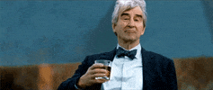 Celebrity gif. Dressed in a tux, actor Sam Waterson raises a glass in a cheers, looking sincere as the wind blows through his hair.
