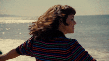 Celebrity gif. Norah Jones outstretches her arms and spins in a circle on the shore before turning to face us.