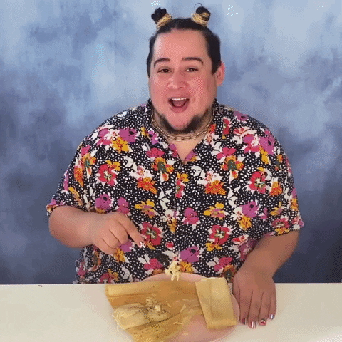 Video gif. Man in a floral shirt and two small high buns in his hair holds a fork over a half-eaten tamale, blowing a kiss at us in a flirtatious way. Text, in Spanish, "Besitos y tamalitos."