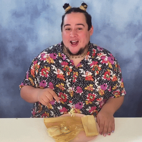 Video gif. Man in a floral shirt and two small high buns in his hair holds a fork over a half-eaten tamale, blowing a kiss at us in a flirtatious way. Text, in Spanish, "Besitos y tamalitos."