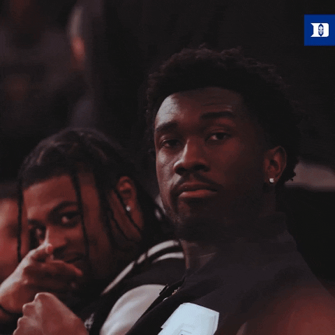 Sports gif. Trevor Keels and Mark Williams of Duke basketball are sitting in the audience at a game. They're both eating popcorn and watching the game intently. They realize the camera is on them and smile. Williams gives us a rock on hand shake while shaking the popcorn box.