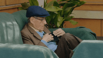 Movie gif. An elderly Sergio Chamy in The Mole Agent sits on a couch, fumbling with his cell phone.