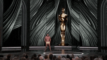 Oscars 2024 GIF. Wide shot of a naked John Cena shuffling to the microphone at the Oscars. He's completely nude, save for some sandals and the Oscar winner envelope covering his precious parts. The giant Oscar statue towers behind him.