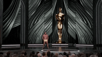 Oscars 2024 GIF. Wide shot of a naked John Cena shuffling to the microphone at the Oscars. He's completely nude, save for some sandals and the Oscar winner envelope covering his precious parts. The giant Oscar statue towers behind him.