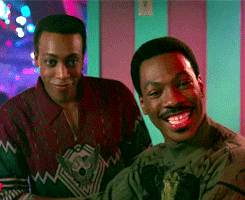 Movie gif. Eddie Murphy as Randy and Arsenio Hall as Semmi in Coming to America. They're at a club and they looking at someone eagerly, with wide grins filling their faces as they lean in to hear better.