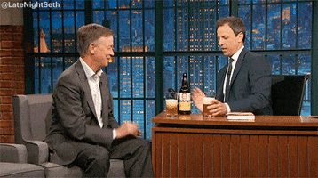Late night show gif. Seth Meyers holds up a glass of beer and clinks it with the man’s cup he’s interviewing.