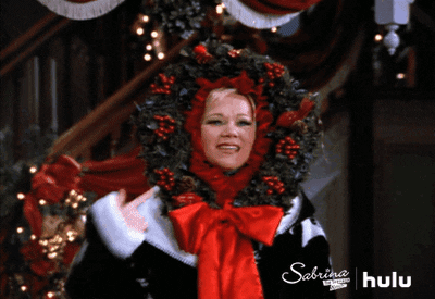 TV gif. Caroline Rhea as Hilda in Sabrina the Teenage Witch wears a Christmas wreath around her head. She touches it and it glows with twinkling lights.