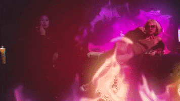 Video gif. A woman reclines on top of a piano and eats casually as colorful flames envelope the space around her. 