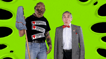 Video gif. Person dressed as the Toxic Avenger holds a mop and bobs its head and points at us, looking back and forth to a senior man dressed like a dorky professor who looks amazed, also pointing at us. Text, "Woah, woah, Toxie look!"