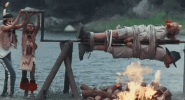 Movie gif. From Addams Family Values, two young campers dressed as Native Americans spin Peter MacNicol as Gary and Christine Baranski as Becky tied-up and gagged on a spit over a campfire.