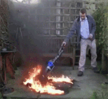 Video gif. Man calmly vacuums over an area of ground that is on fire in a backyard.
