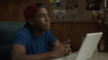TV gif. Jharrel Jerome as Jerome in Mr. Mercedes sits at a table in front of a laptop and folds his hands together while looking up and smiling innocently.