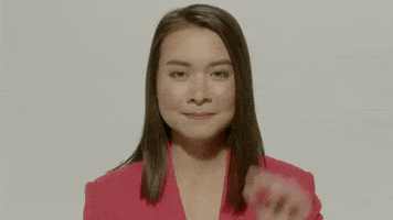 Celebrity gif. Mitski smiles coyly, holds her hand up and waves at us.