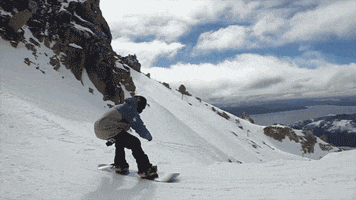 Sports gif. Powder flies as a snowboarder makes a jump into the air, holding his board and spinning as he lands on the side of a mountain.