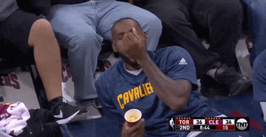 Sports gif. Lebron James sits on the bench with the Cleveland Cavaliers, chilling and eating some popcorn out of a cup. 