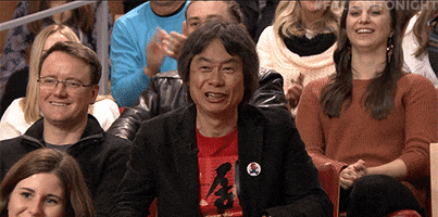 Celebrity gif. Shigeru Miyamoto, in TV audience, extends a thumbs down and frowns, but then turns it into a thumbs up and laughs as the crowd laughs and cheers around him.
