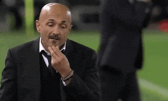 Sports gif. Luciano Spalletti on a soccer pitch looks down unhappily as he strokes his chin and walks towards us. He grimaces slightly, just showing his teeth. 