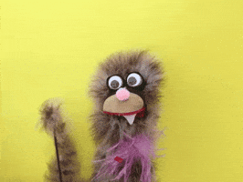 Video gif. A fuzzy puppet with different shades of brown hair, has large bobble eyes. One pointed tooth sticks out from under its tan snout. A stick holds up one of the puppets hands. It's long arm waves quickly. 