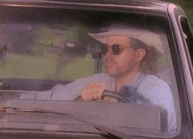 Music video gif. From the video for "Who's That Man," Toby Keith sits behind the wheel of a truck, wearing sunglasses and a cowboy hat, putting his arm around the passenger seat headrest and looking through the windshield.