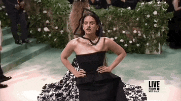 Met Gala 2024 gif. Rosalia poses with hands on hips wearing a black Dior Haute Couture strapless gown with a straight neckline that flows into a train that cascades from her hips and out behind her. She wears a netted square-shaped veil that sweeps down across her face.