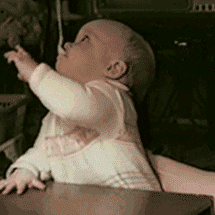 Video gif. A baby is holding a spoon in its mouth and they're trying to reach for it but failing, not understanding how the spoon is staying upright in its mouth. They've just discovered their lip muscles.
