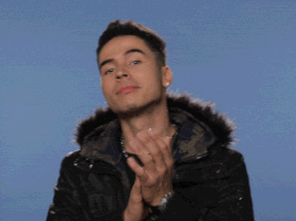 Celebrity gif. Reykon claps and nods approvingly, wearing a heavy black jacket with a fur hood against a steel blue background.