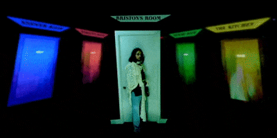 Music video gif. Briston Maroney stands in the middle of a dark environment with rows of colorful doorways to his left and right.