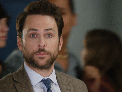Movie gif. Wearing a brown suit jacket, Charlie Day gives us a blank look as several people walk by him in the background. After a second, he raises a hand to give us a thumbs up, then walks out of frame.