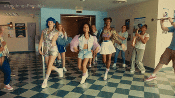 Movie gif. A Scene from Anything’s Possible. Eva Reign as Kelsa dances with her two best friends as cheerleaders jump up with pom pom behind them. Band kids play their instruments around them in the middle of a school hallway. 