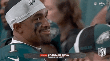 Sports gif. Brandon Brooks of the Philadelphia Eagles sits on the sidelines wearing his uniform and a white headband. He's looks over his shoulder and grins from ear to ear in slow motion. 