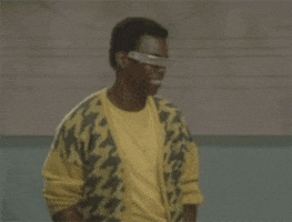 Video gif. Man wearing a patterned banana yellow and dark green sweater vest along with an eye shield that completely obscures his eyes gives us a flashy smile and a thumbs up.