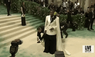 Met Gala 2024 gif. Colman Domingo wears an oversized, white-and-black Willy Chavarria suit with diamond earrings and a bouquet of white flowers wrapped in a white satin cloth. This is paired with a full-length ivory colored cape that hangs from the shoulders and sweeps back in a short train. 
