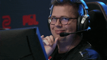 Sports gif. FaZe Karrigan sits in a chair with headphones on at an esports event and waves at us, wiggling his eyebrows and blowing us a kiss as we zoom in.