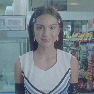 Music video gif. Olivia Rodrigo in her video for good 4 u. She stares at the camera and shrugs sweetly while wearing a cheerleading uniform.