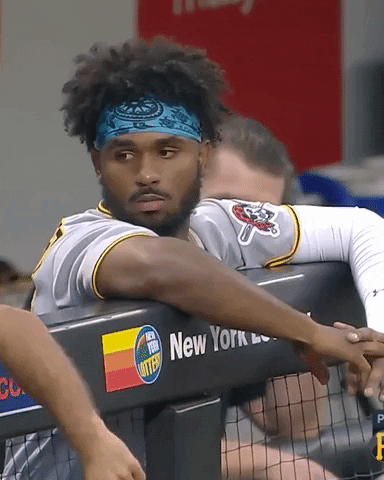 Sports gif. Gregory Polanco of the Pittsburgh Pirates stands behind a net in the dugout as he looks over and casually fist bumps someone standing next to him. 