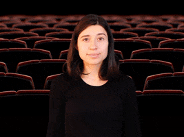 Digital compilation gif. Young woman claps her hands in an exaggerated way then leans forward at us as popcorn pops up into the air all around her in front of a movie theater background. Text, "The movie was better."
