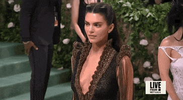 Met Gala 2024 gif. Kendall Jenner wearing a plunging black and bronze vintage Givenchy 1999 Haute Couture gown, shifts shyly, lost in her mind.
