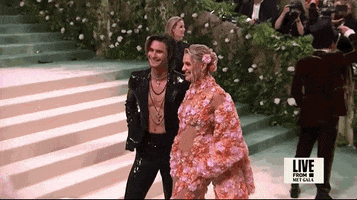 Met Gala 2024 gif. We zoom in on Kelsea Ballerini and Chase Stokes posing together. Ballerini is wearing a Michael Kors dress with vibrant floral appliqués accented by irregular sheer panels across the torso. She has a thick matching shrug covered in matching floral appliqués and flowing cape sleeves. Stokes is wearing a Michael Kors black suit covered in black sequins and a low-cut open blazer with no shirt, layered necklaces, and a wet hair look.