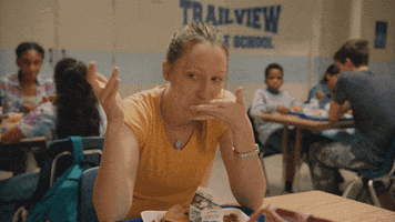 TV gif. Anna Konkle, as Anna on Pen15, sits at a cafeteria table with a tray of food, raising her eyebrows and smiling as she licks her fingers.
