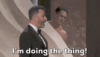 Oscars 2024 GIF. A potentially nude John Cena is behind a wall on stage and he is totally stressed out. Jimmy Kimmel stands in front of him and Cena gestures with his face for Kimmel to come to him now. Kimmel says, "I'm doing the thing!" but Cena insists. 