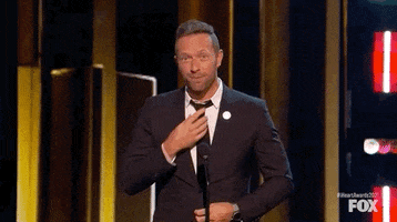 Celebrity gif. Chris Martin at I Heart Radio Music Awards adjusts his tie nervously, crosses his fingers, and says "fingers crossed."