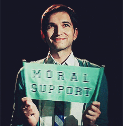 Video gif. A man with a modest smile waves a small collegiate-style banner that says, "Moral support."