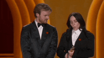 Oscars 2024 gif. Billie Eilish and Finneas O'Connell win Best Song. O'Connell looks at Eilish while she sucks in her cheeks before bending down and breaking into laughter. 