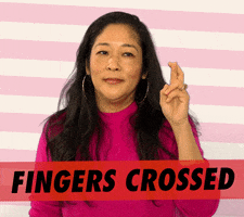 Video gif. A woman wearing a magenta sweater and hoop earrings holds up crossed fingers. She speaks with a slight eye roll, as if she's not particularly hopeful for this outcome. Text, "Fingers crossed."