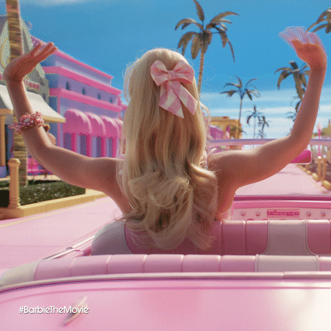 Movie gif. Margot Robbie as Barbie drives a pink convertible through the driveway past a pink movie theater, looking very friendly as she smiles and waves with both hands at everyone she's passing on the street.