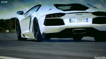 Video gif. From behind, a white Lamborghini Aventador drifts gently on the road before catching traction and turning.