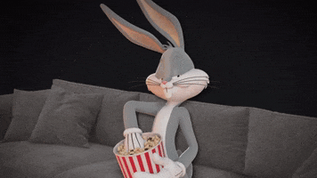Looney Tunes gif. Bugs Bunny takes a handful from a striped bucket full of popcorn and tosses it into his mouth, looking at us slyly while sitting on a couch.