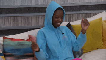 Reality TV gif. A contestant from Big Brother is sitting on the couch wearing a hoodie with the hood on and she does a little dance with her arms.